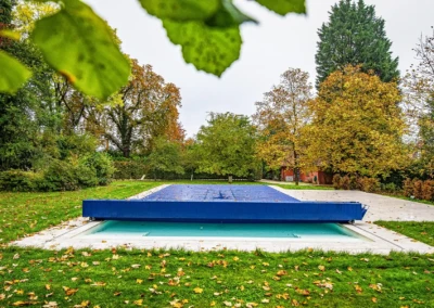 Une piscine couverte par une bâche bleue est entourée de pelouse verte et de grands arbres aux feuilles d'automne.