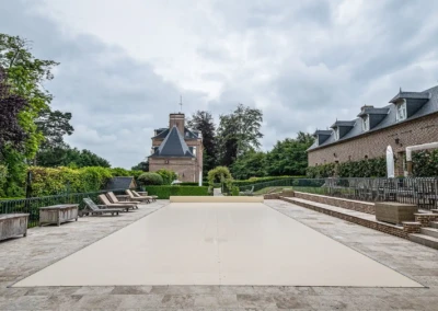 Piscine couverte de beige entourée de chaises longues, située devant un bâtiment en briques avec un ciel nuageux.