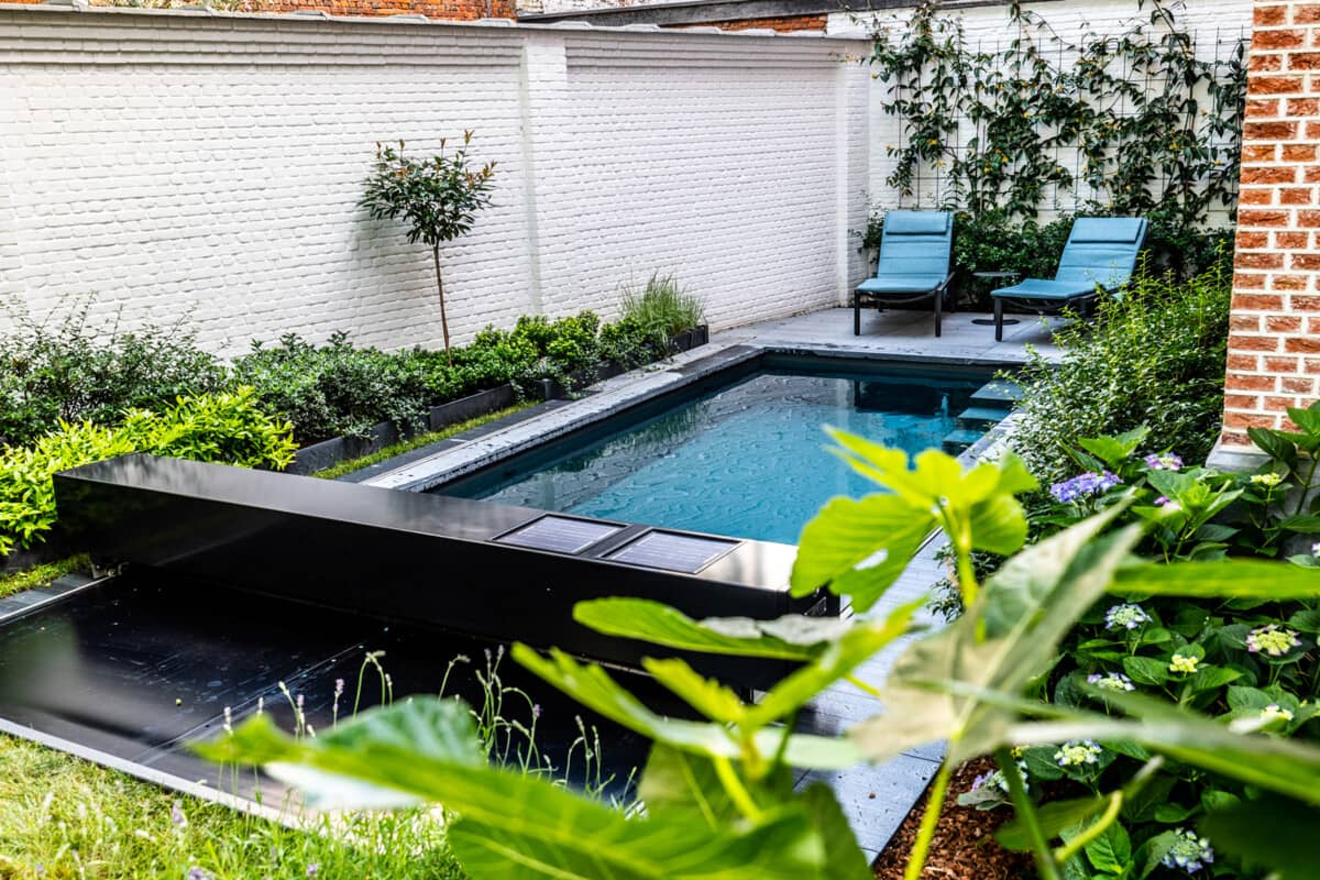 Petite piscine rectangulaire entourée de végétation verte avec deux chaises longues bleues sur une terrasse en pierre grise.