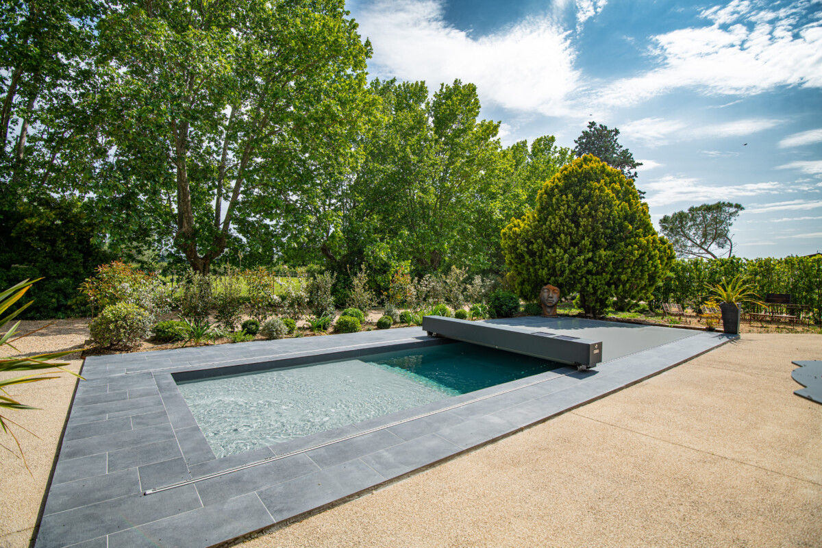 Piscine rectangulaire entourée de dalles grises, avec un volet de protection, bordée de végétation luxuriante sous un ciel partiellement nuageux.