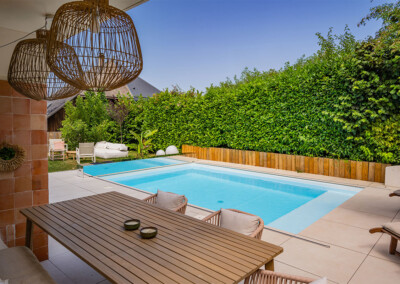 Vue depuis une terrasse couverte donnant sur une piscine équipée d’une Coverseal, entourée de haies végétales et d’un mobilier de jardin naturel.